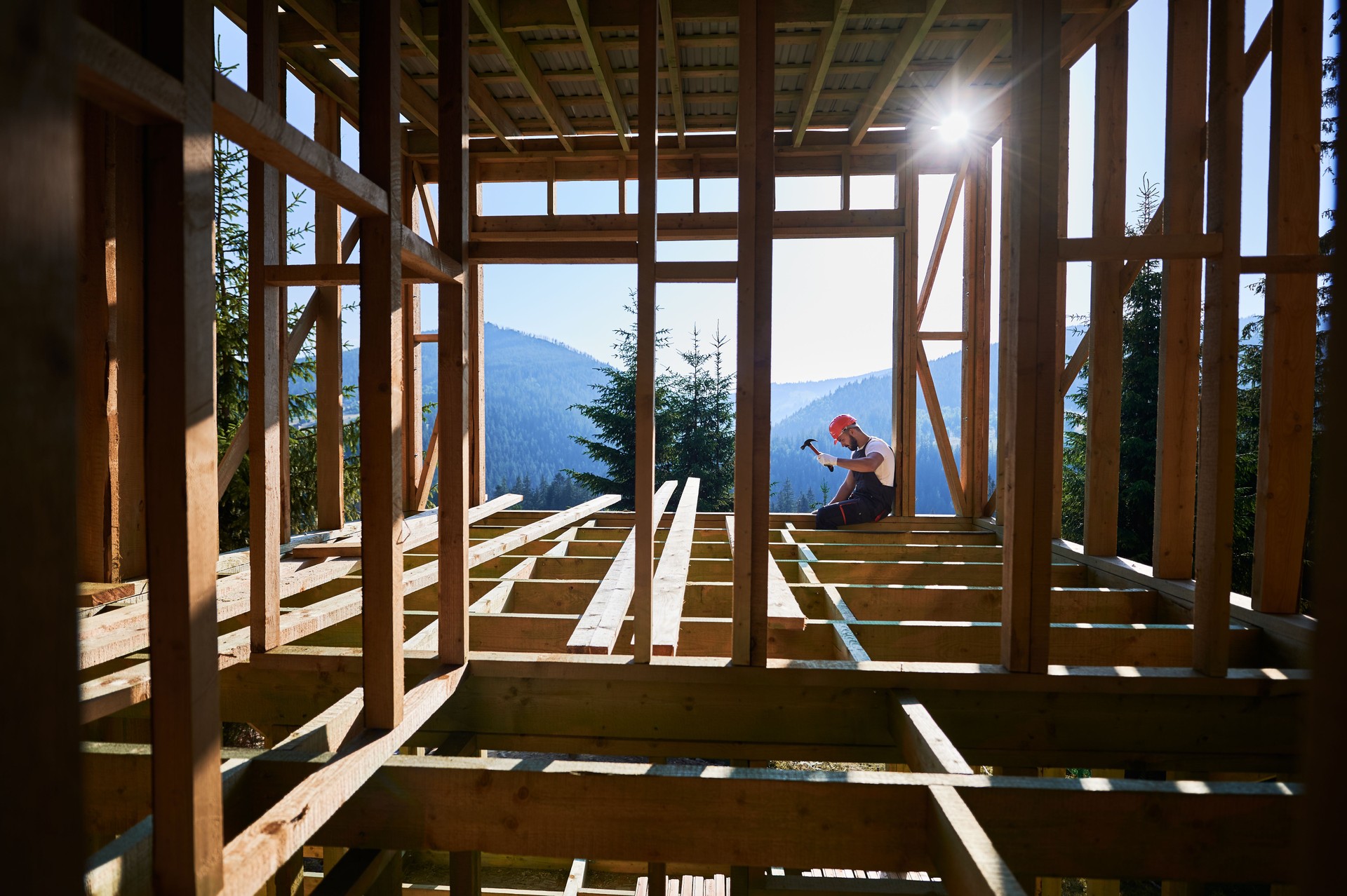 Carpenter hammering nail while constructing wooden frame two-story house near the forest.